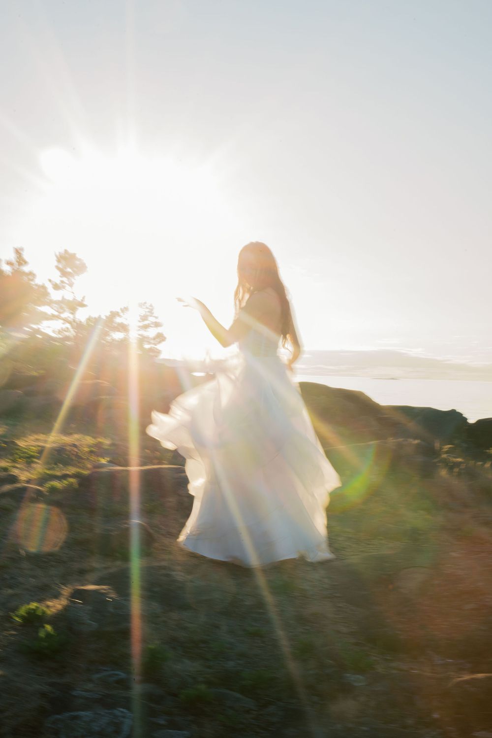 Bride in flowing white gown standing on rocky cliffs with sun flares and ocean views in background.