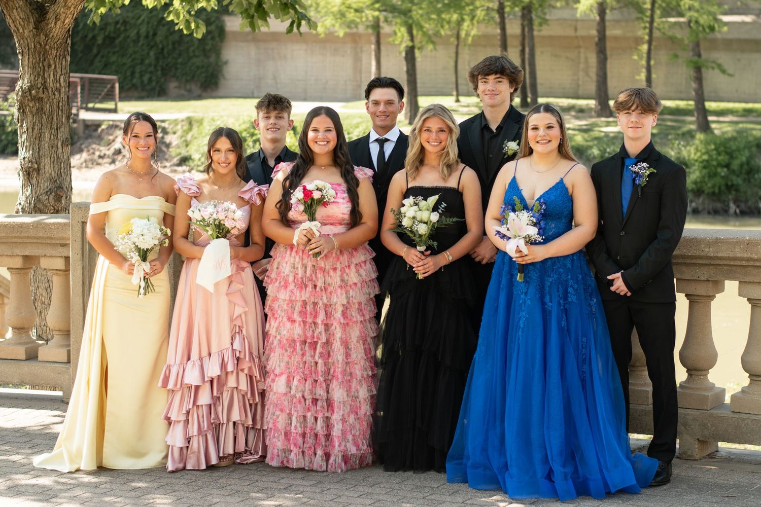 A bridal party in mixed formal dresses poses together on a lush lawn by a lake.