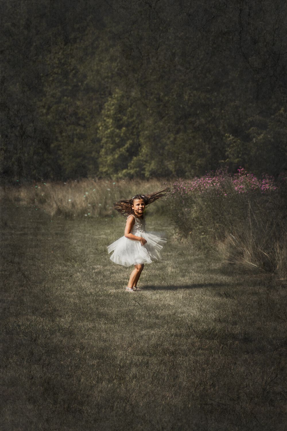 Child twirling in a white dress against a moody, dark landscape with flowering bushes.