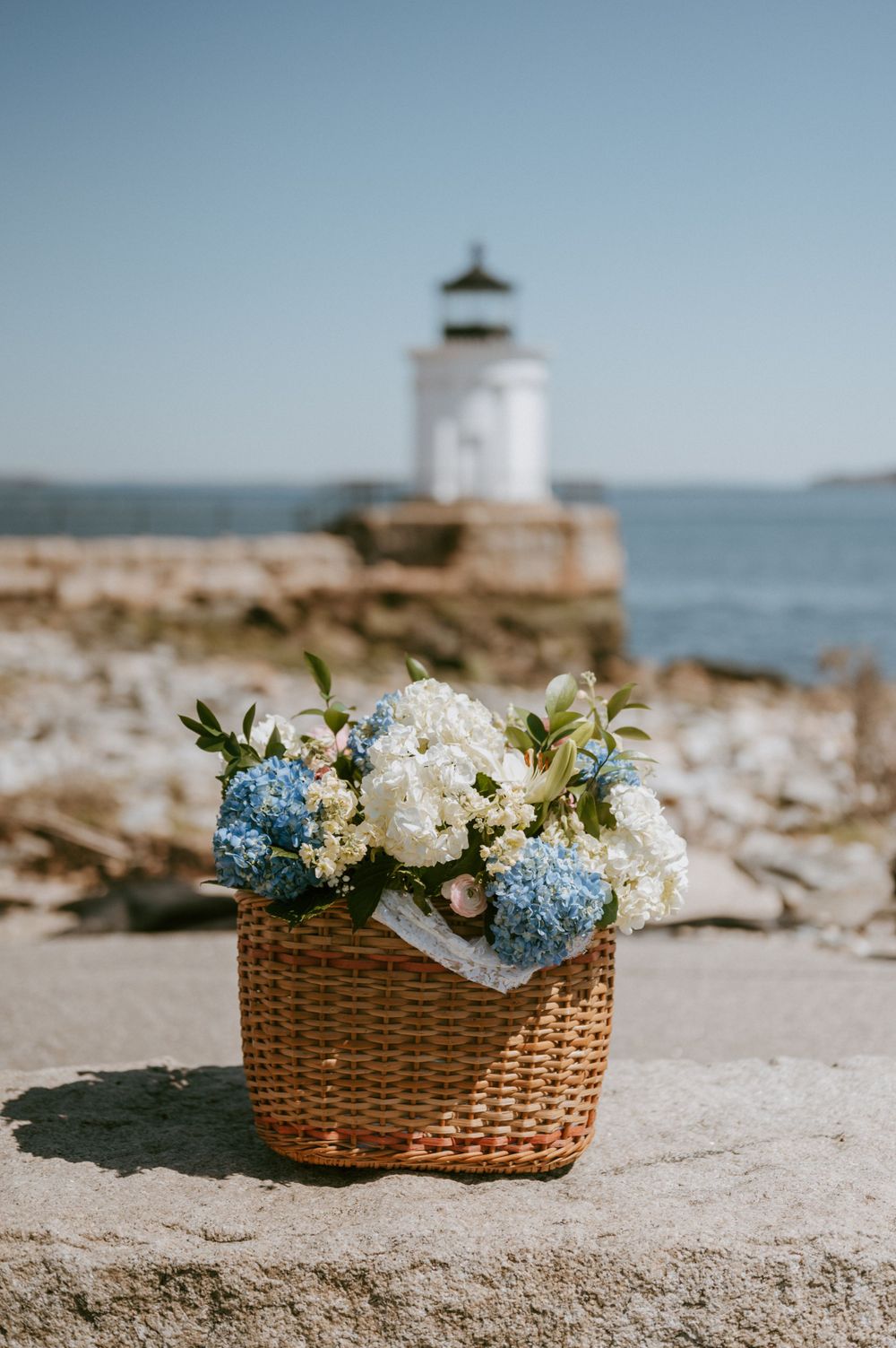 Woven basket filled with blue and white hydrangeas sits near a lighthouse on a rocky coastal shore.