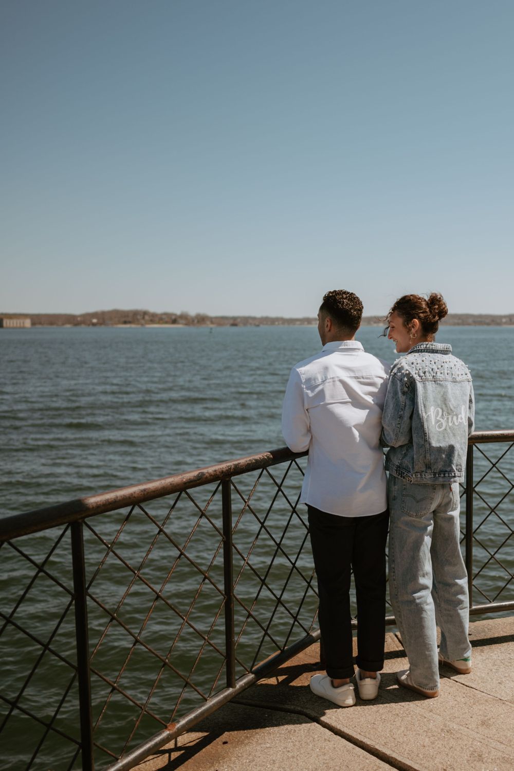 Two figures lean against railing overlooking waterfront on sunny day.