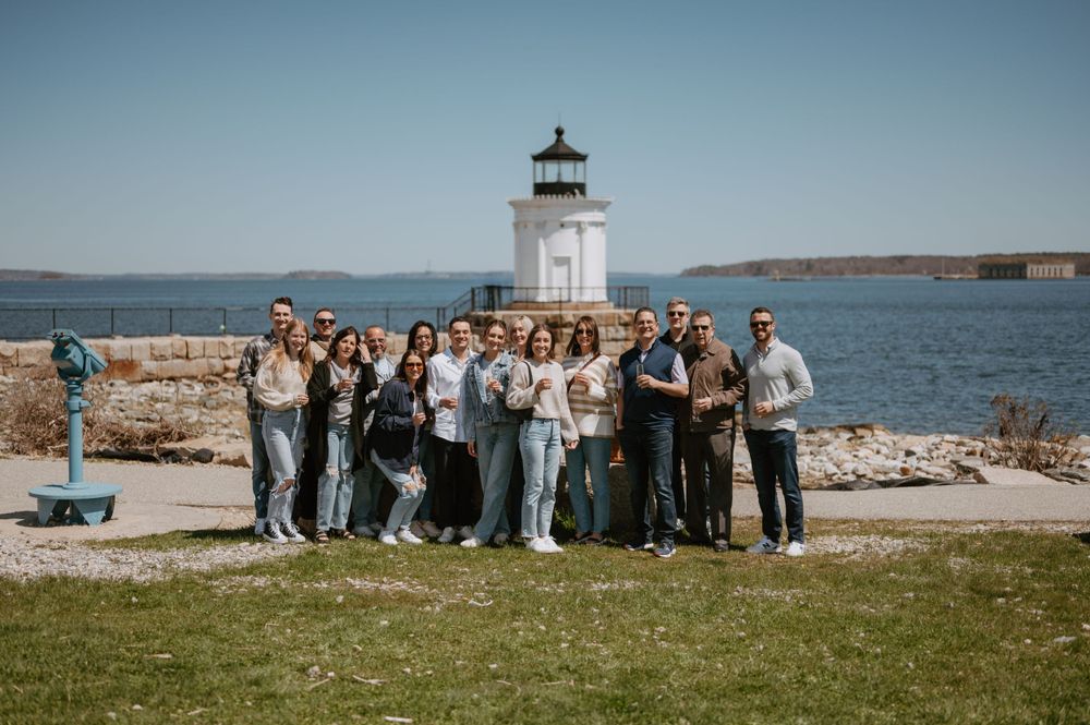 Group photo of wedding attendees gathered in front of a coastal lighthouse.