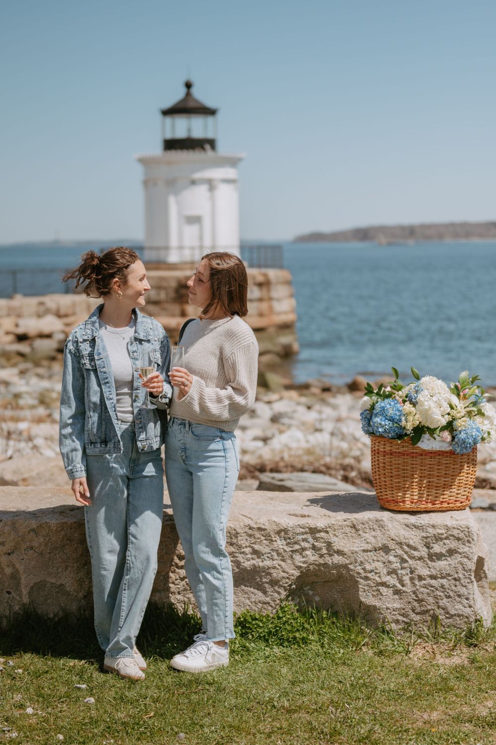 Two people share drinks by lighthouse with flower basket on stone wall.