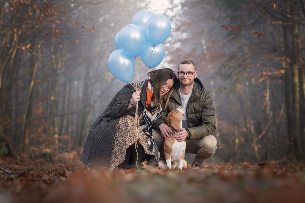 Two people and their dog posing with blue balloons in an autumn forest setting.