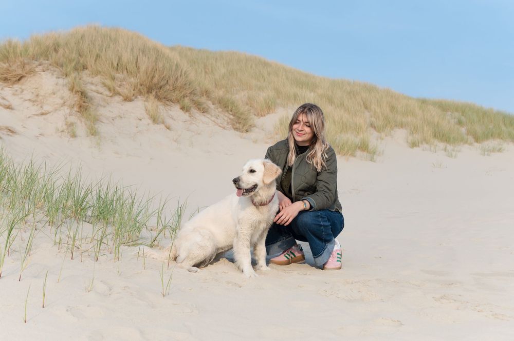 A person in a denim outfit sits with their white golden retriever dog on a sandy beach dune with tall grass.