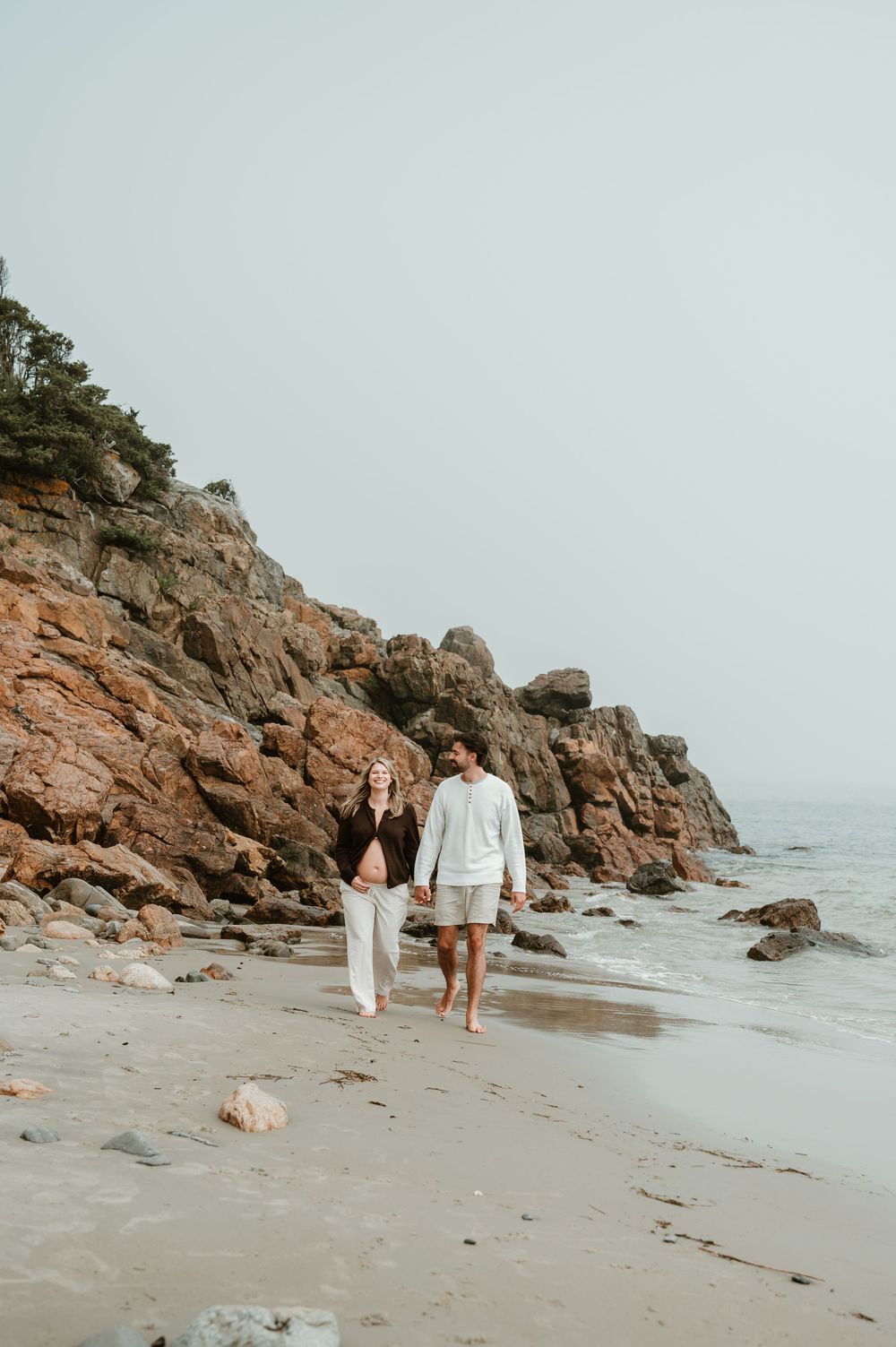 A couple walks hand-in-hand along a rocky beach shoreline, with red cliffs and trees in the background.