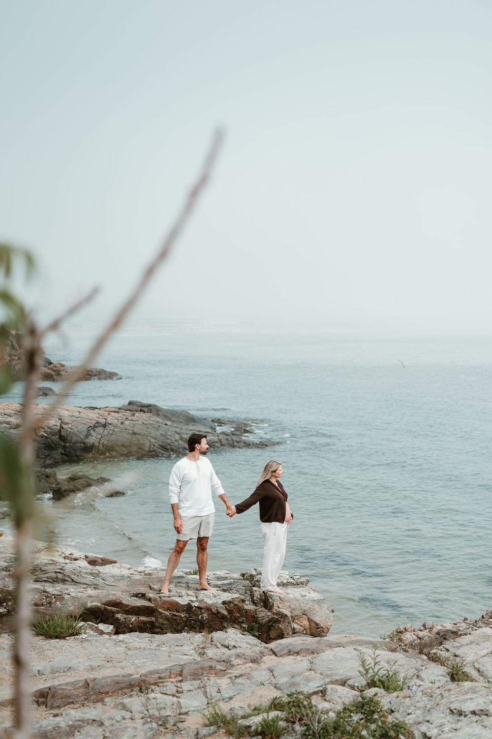 Couple holding hands on rocky coastal shoreline, man in white outfit and woman in dark top with white pants, ocean backdrop.