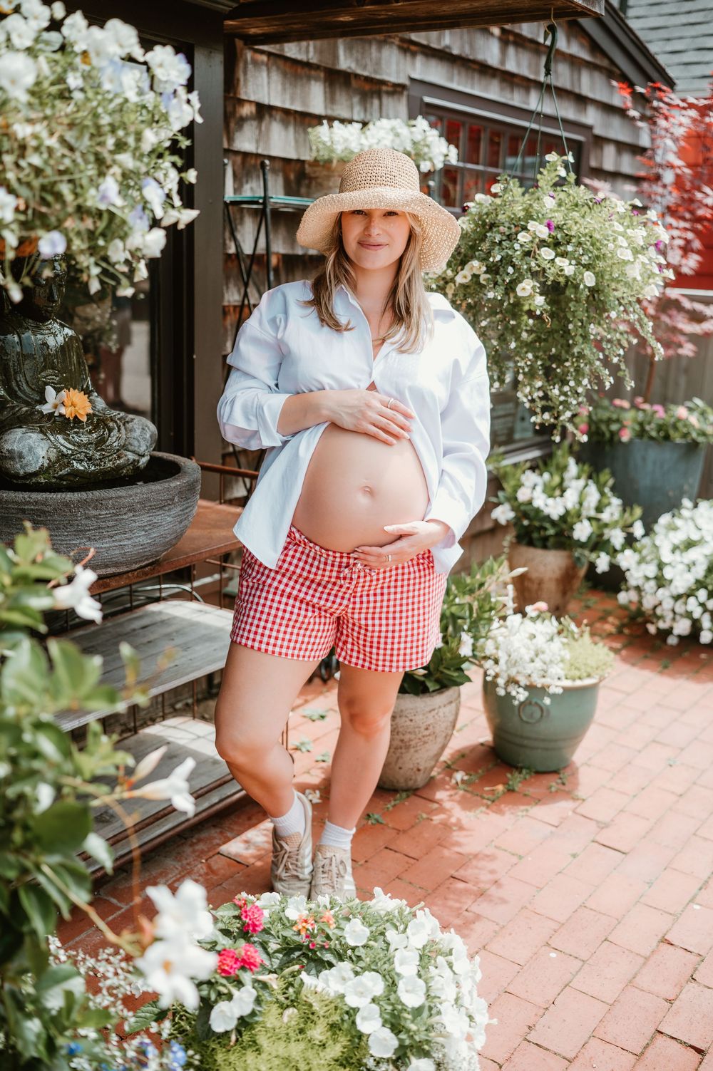 Pregnant woman in straw hat, white shirt, and red gingham shorts posing outdoors among blooming flowers.