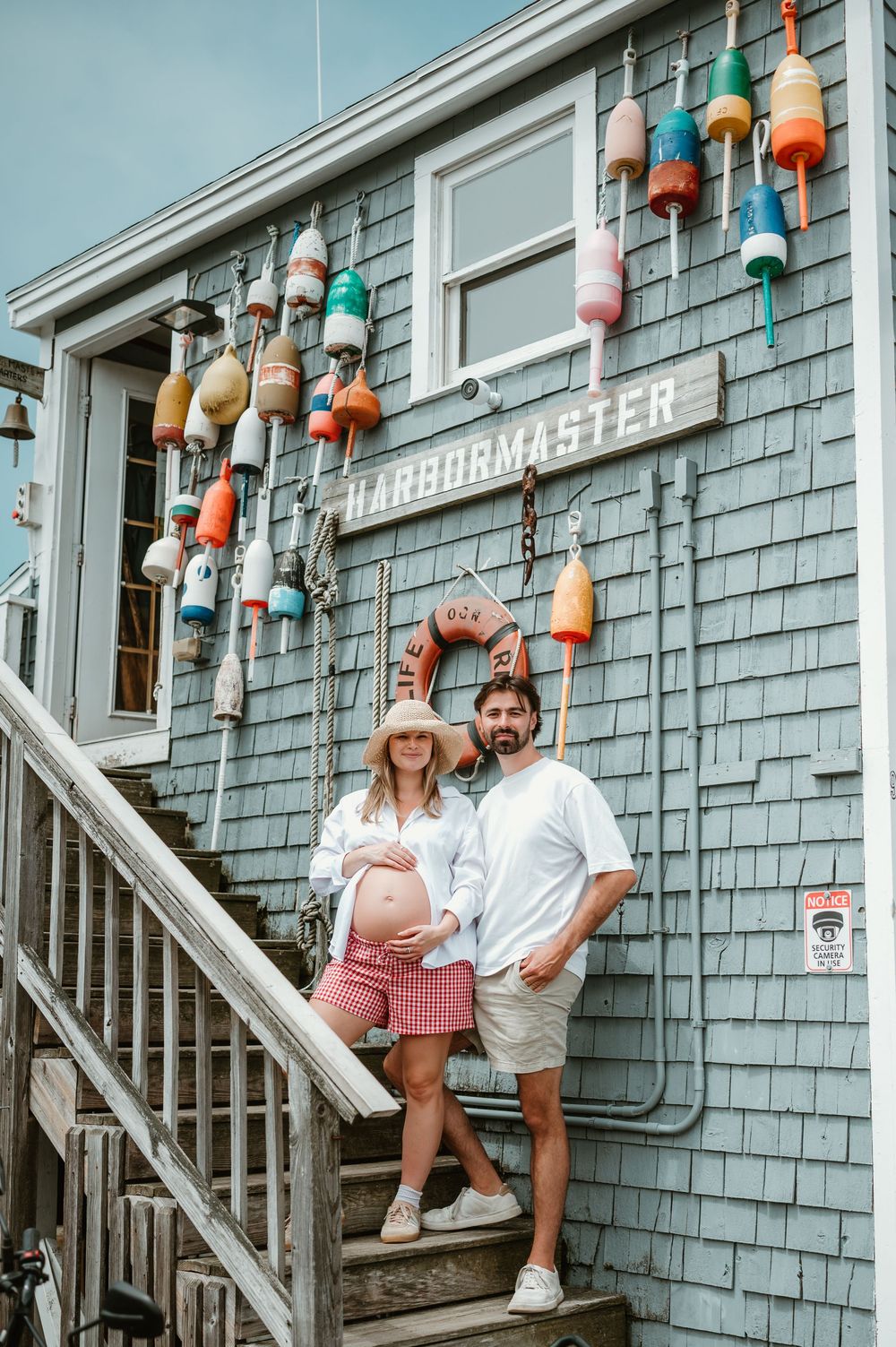 Pregnant couple posing in front of a harbormaster building decorated with colorful buoys on a coastal New England dock.