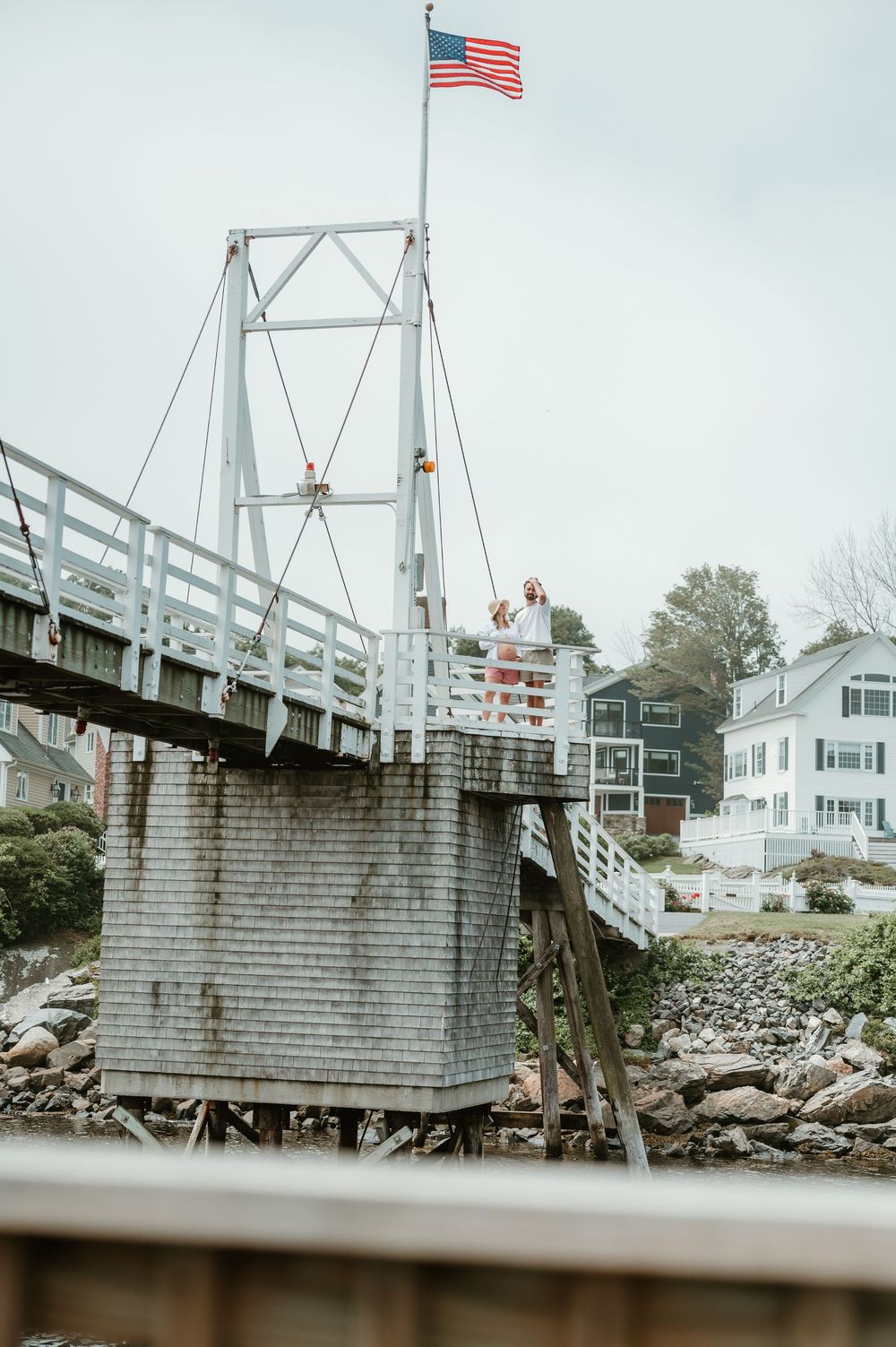 A white drawbridge with an American flag flies over rocky shores near coastal New England homes on a cloudy day.