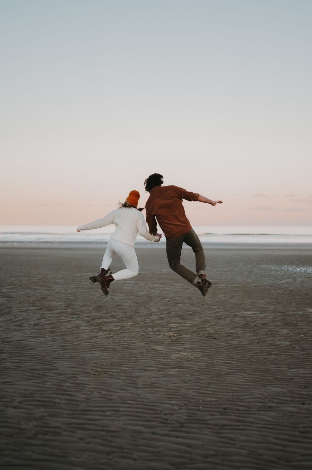Couple jumping together on beach at sunset in playful moment.