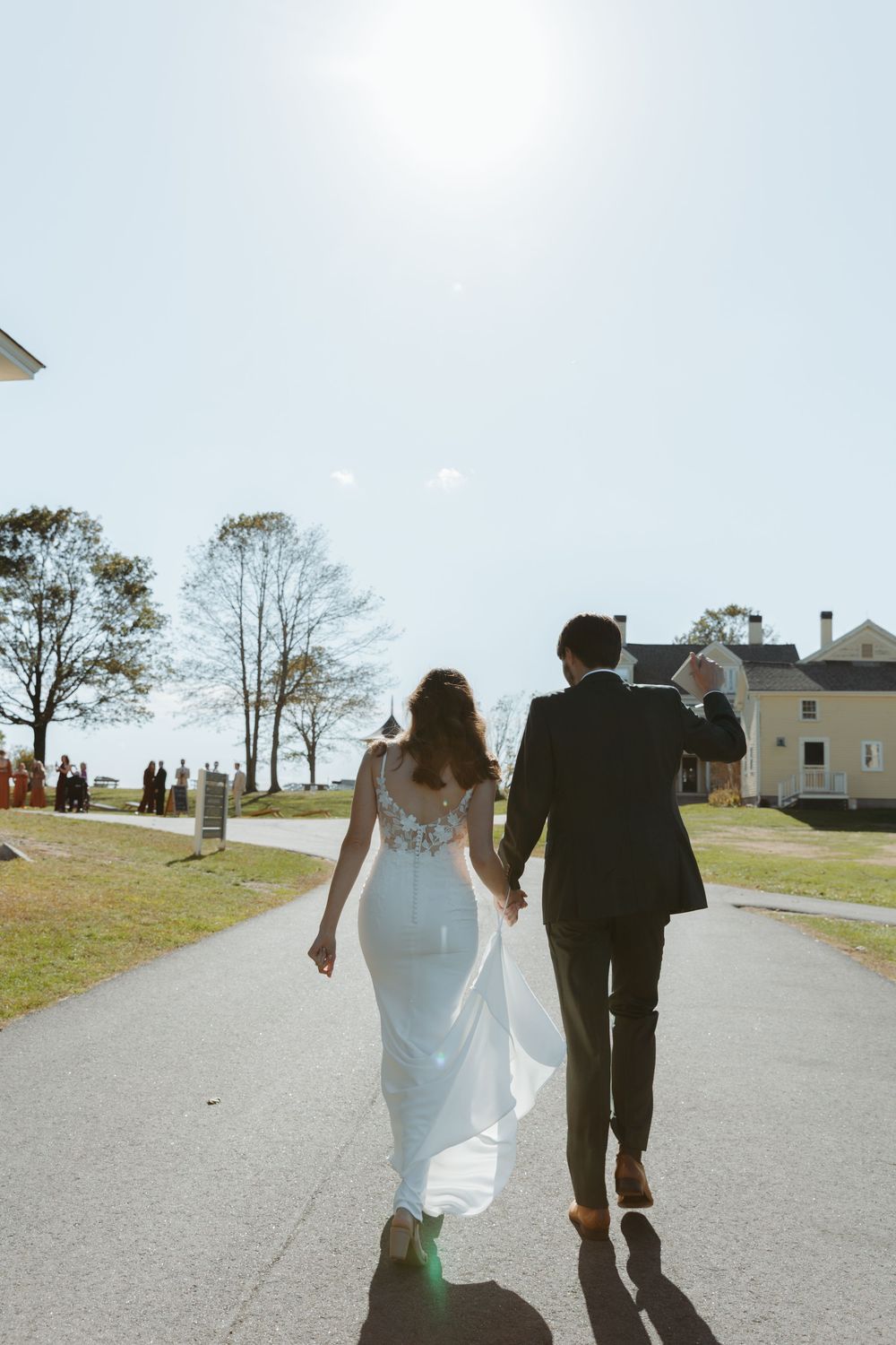 A couple in wedding attire walks away on a sunny path with houses and trees in the background.