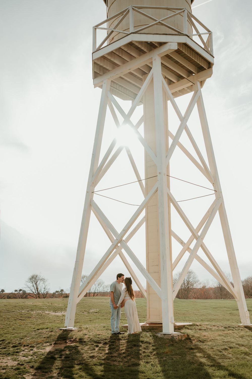Couple sharing a romantic moment under a white water tower in an open green field on a cloudy day.