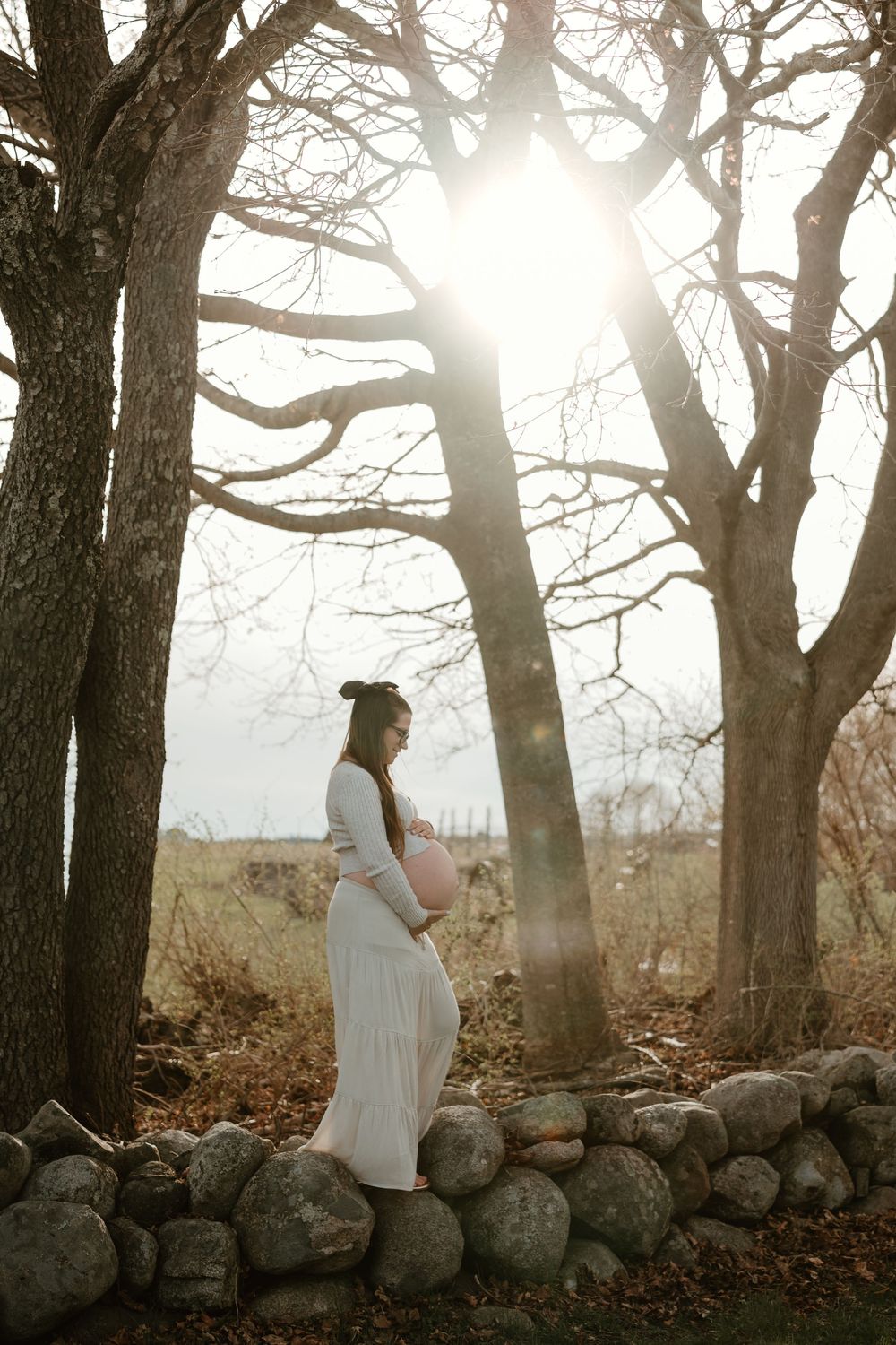 Pregnant woman in white dress cradling her belly outdoors by a stone wall, surrounded by bare trees with sunlight.