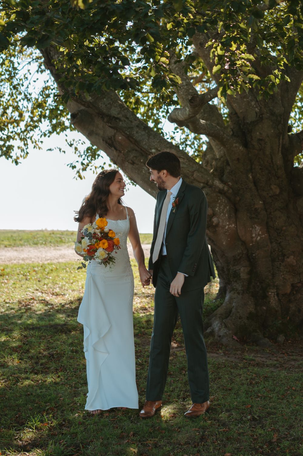 A couple shares a tender moment under a large tree during their outdoor wedding ceremony with golden sunlight filtering through.