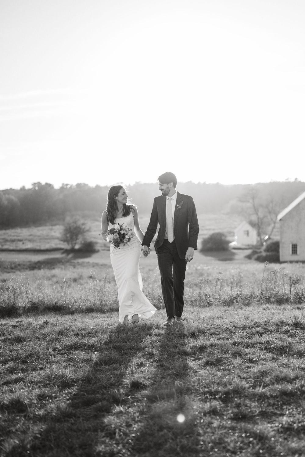 A couple in wedding attire walks through a sunlit meadow in black and white.