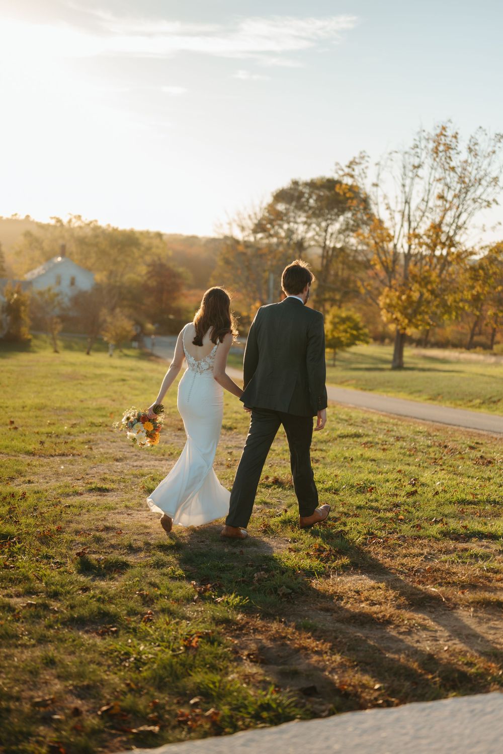 A couple in wedding attire walks hand in hand along a sunlit path during golden hour as autumn trees glow in the background.