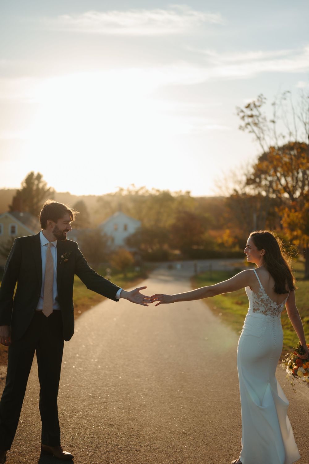 Couple in wedding attire dancing together on a path at sunset with warm autumn light glowing behind them.