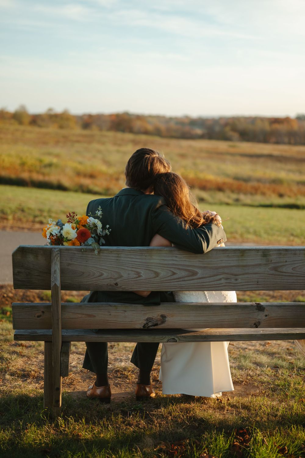 A couple sits together on a wooden bench at sunset in a serene autumn field with a small floral arrangement beside them.