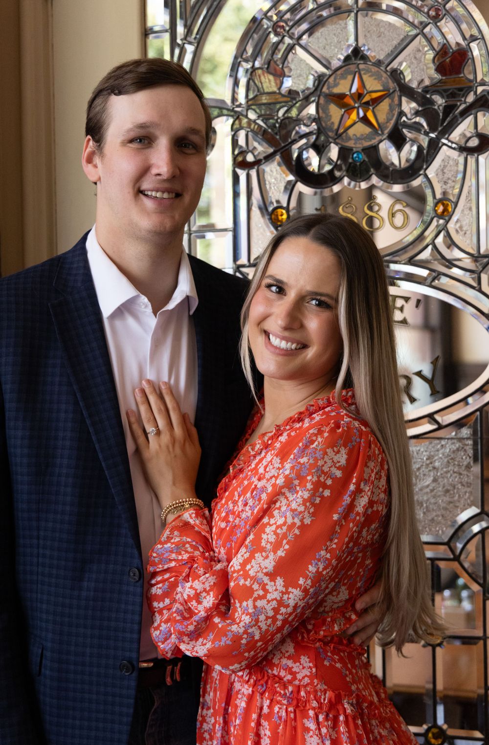 Engaged couple posing by decorative stained glass window in historic building.
