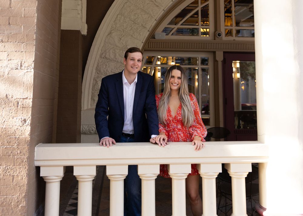 Couple standing on balcony with stone columns and arches in architectural setting.