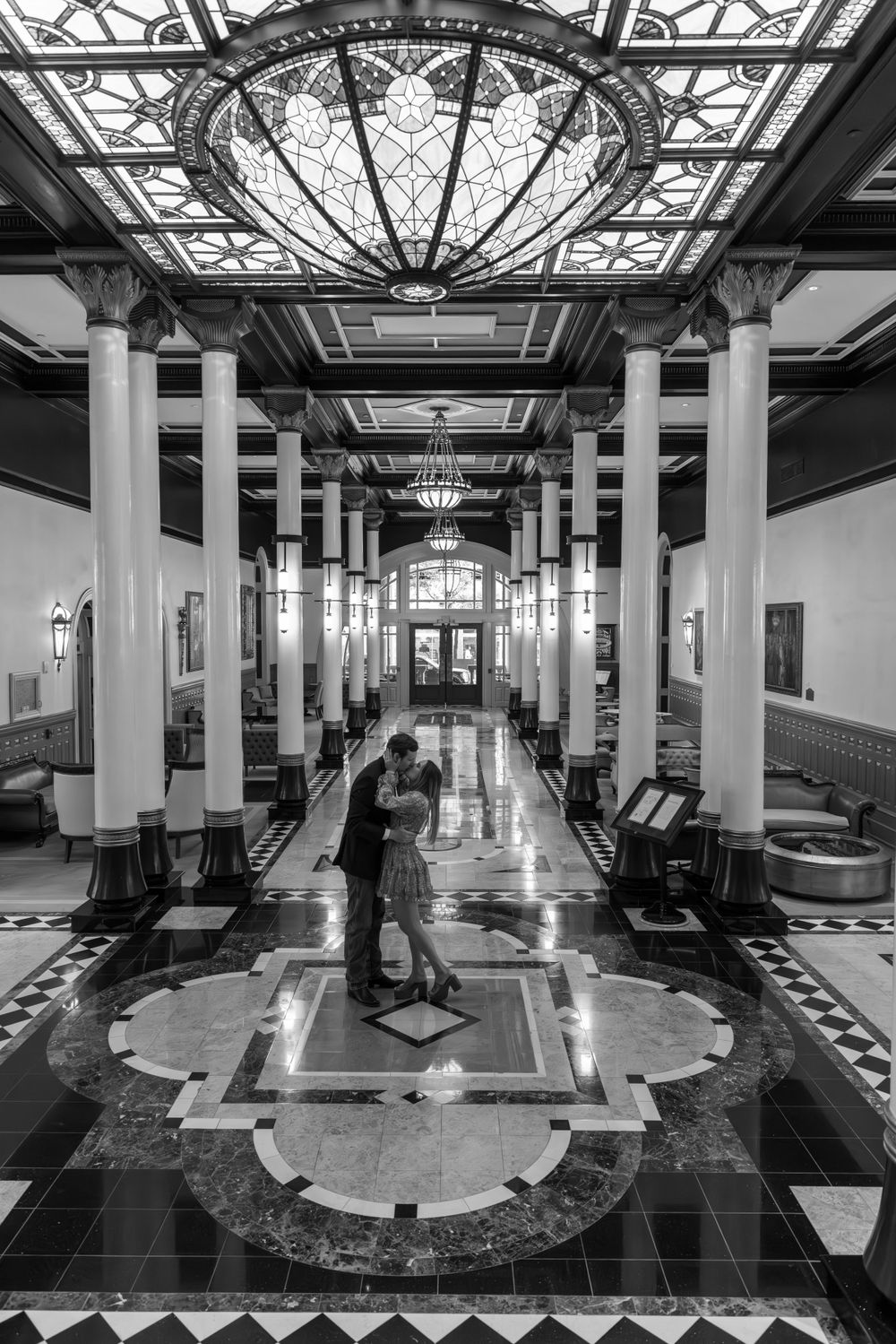 Couple captured in black and white under ornate ceiling with dramatic lighting in grand hallway.