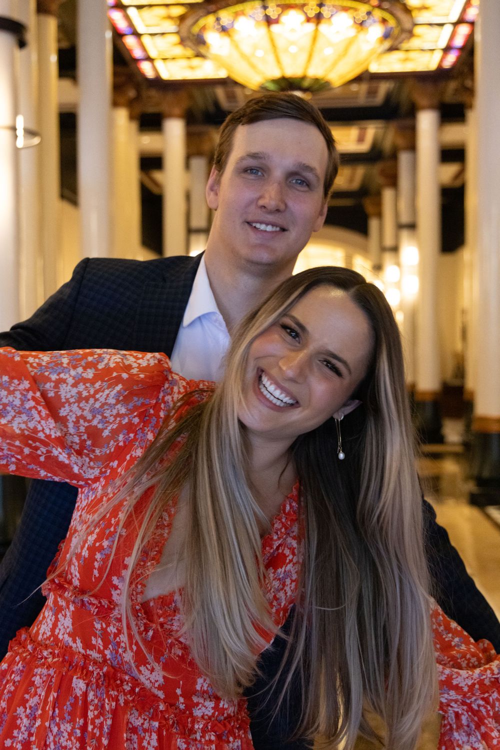 Elegant couple posing beneath vintage chandelier in historic building hallway.