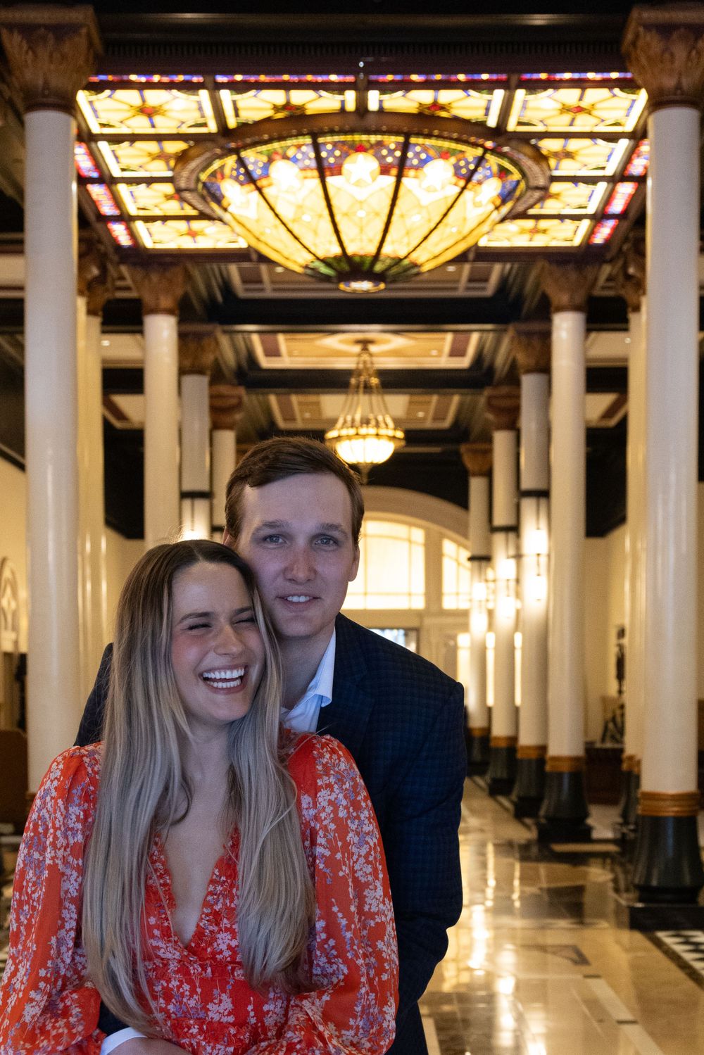 Interior of historic building with ornate stained glass ceiling and elegant lighting fixtures with happy newly engaged couple.