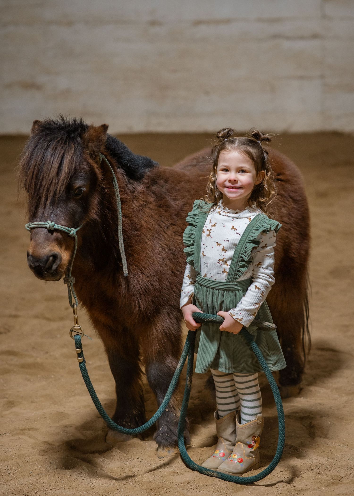 A small brown pony stands beside a child in green overalls inside a riding arena.