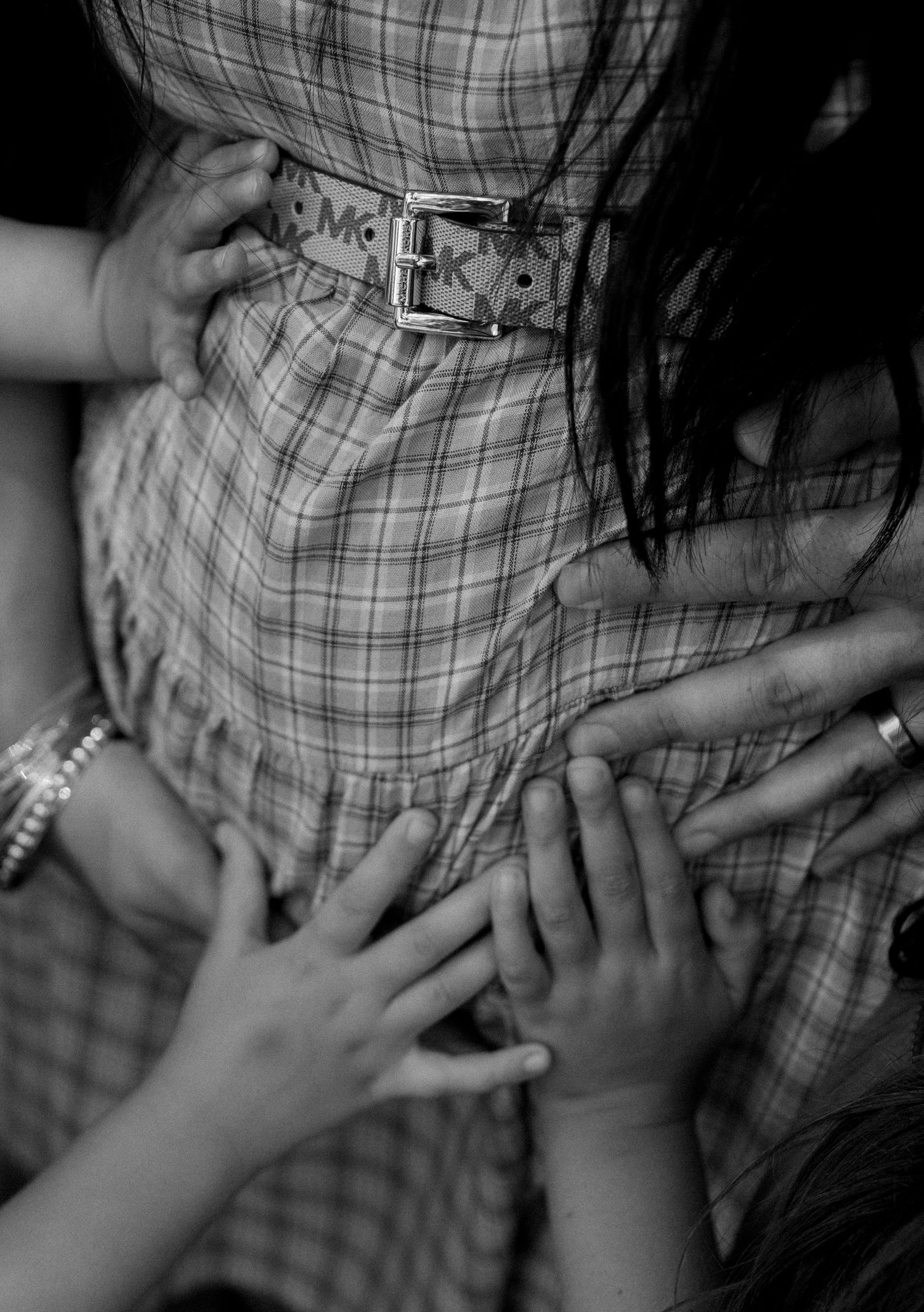 Black and white photo of children's hands touching pregnant woman's belly in plaid dress.