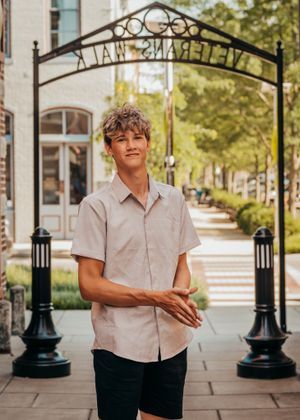Young person in a short-sleeve beige button-down shirt and dark shorts stands under a decorative archway on a brick sidewalk.