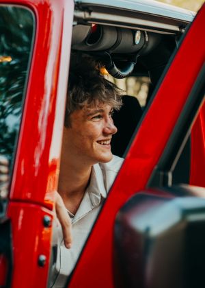 Highschool graduate poses in the open-door front seat of his cherry red jeep with black trim during portrait session.