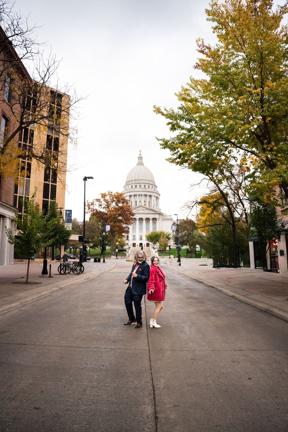 Couple posing on a street in front of the Wisconsin State Capitol building surrounded by autumn trees.
