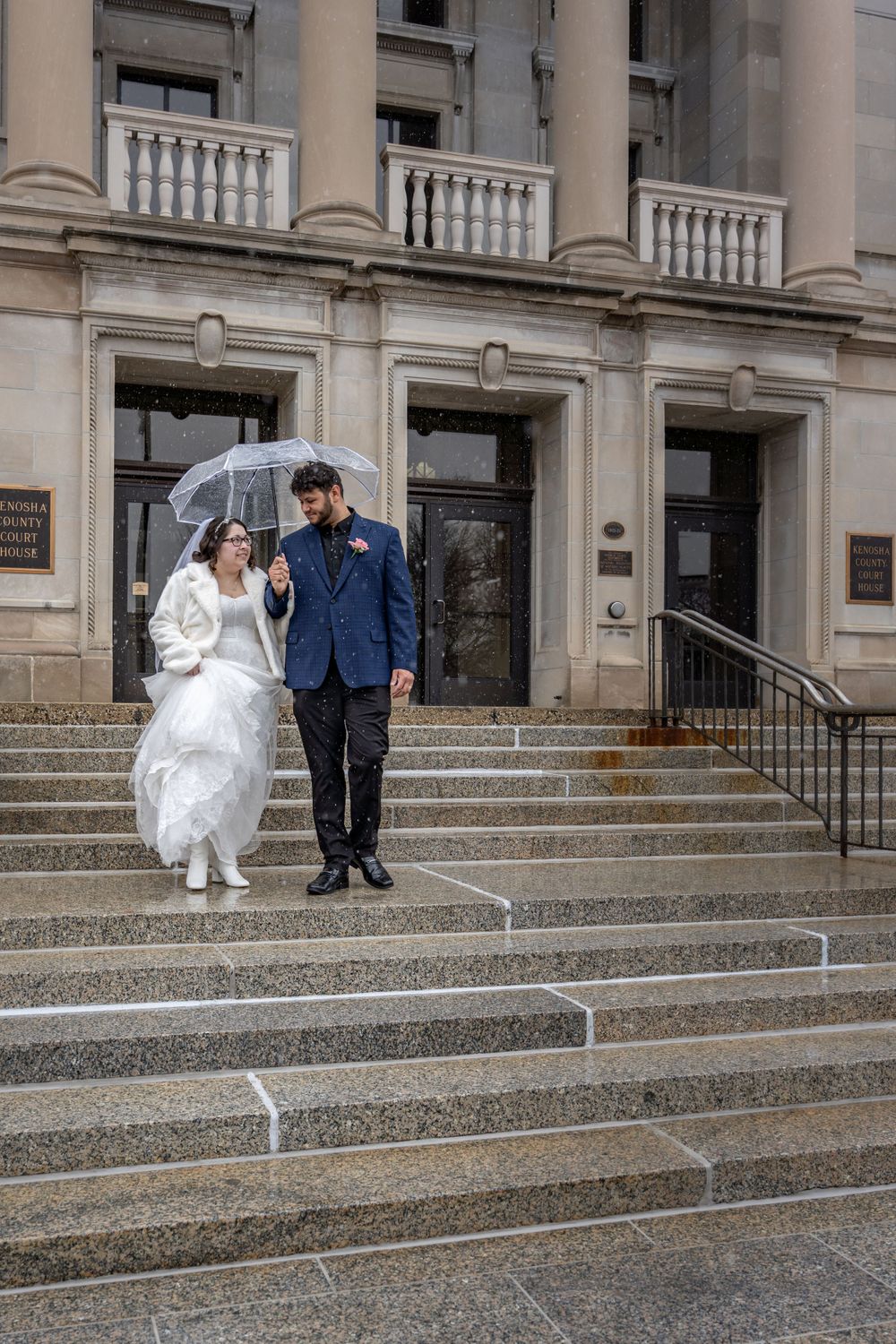 A bride and groom stand on courthouse steps under an umbrella on a rainy wedding day.