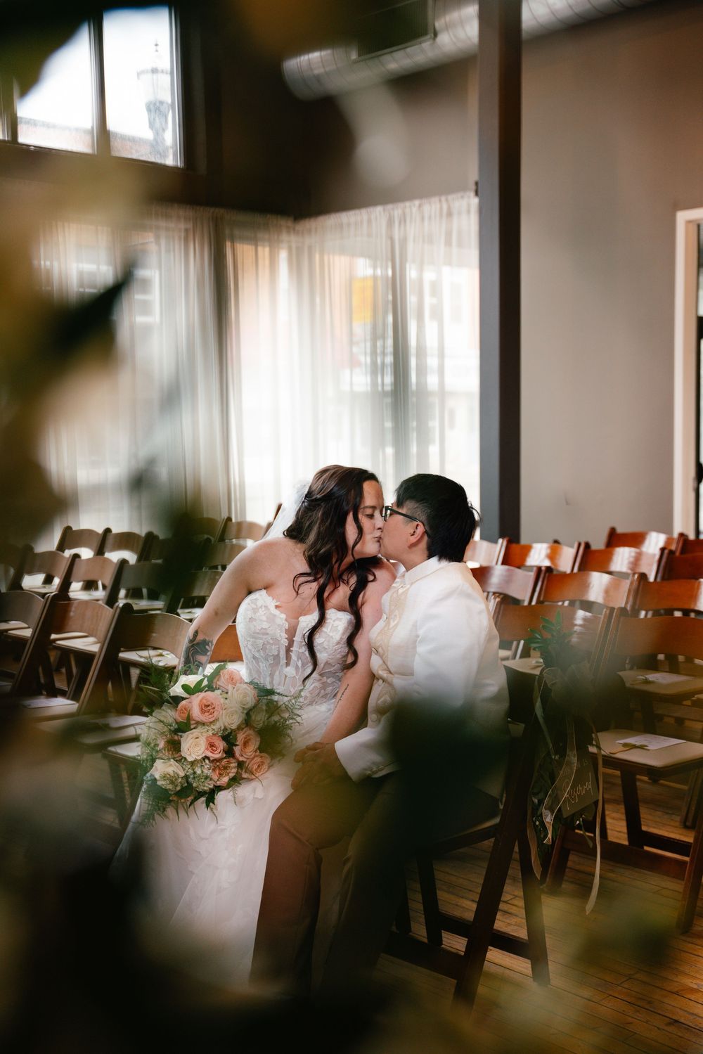 Bride and groom share a romantic kiss while seated among wooden chairs in a warmly lit indoor wedding venue.