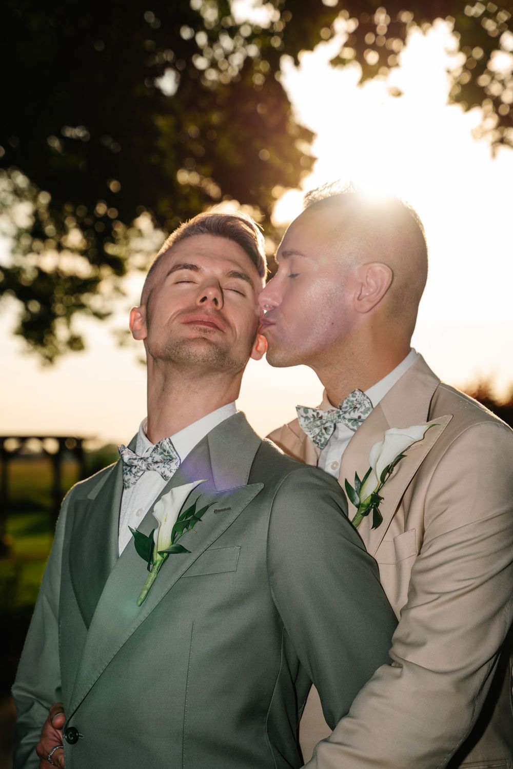 Two grooms in matching bow ties share a romantic kiss at sunset during their outdoor wedding ceremony.