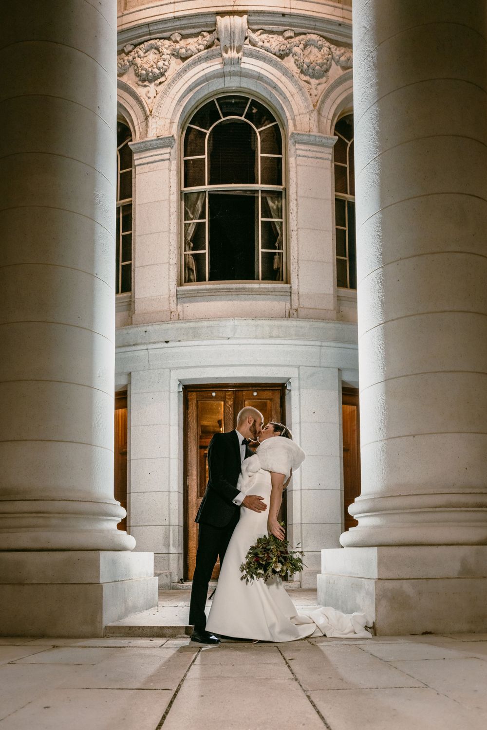 A couple embracing between classical columns at an elegant building entrance.