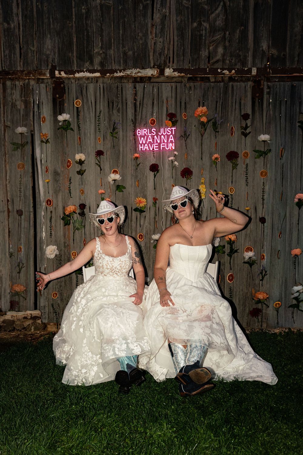 Two brides in white gowns, cowboy hats, and heart sunglasses pose playfully in front of a floral barn backdrop.