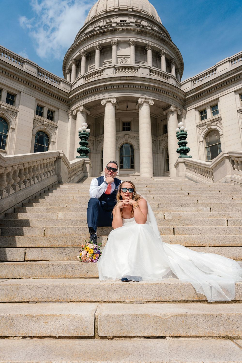 Bride and groom sitting on steps of Rhode Island State Capitol building, wearing sunglasses, with bridal bouquet nearby.