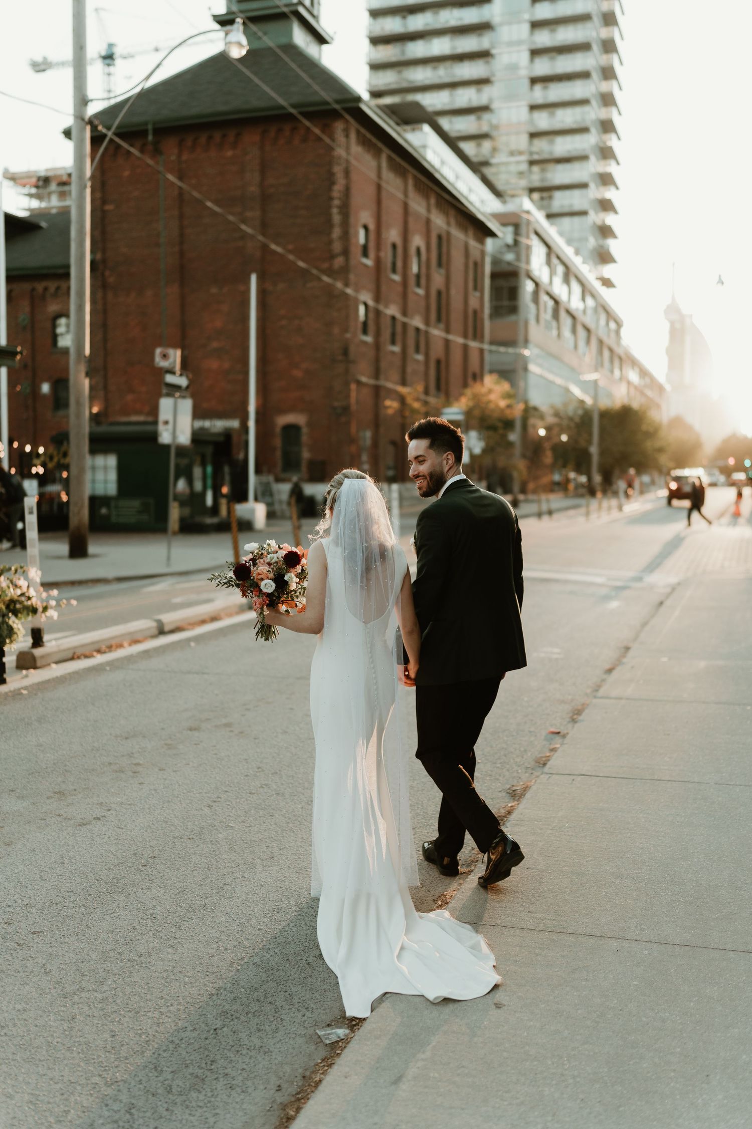 Bride and groom walk down a city street at sunset with urban architecture in background.