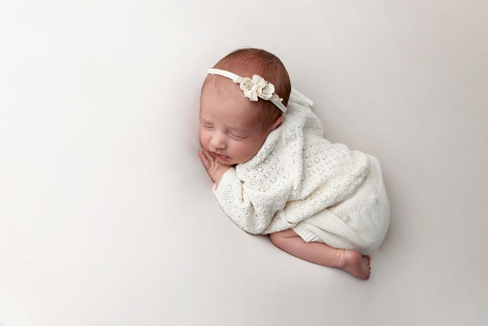 A sleeping newborn baby wearing white lies peacefully on a soft white blanket during a professional photo session.