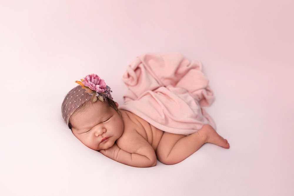 Newborn wearing pink headband peacefully sleeping on rose-colored fabric.