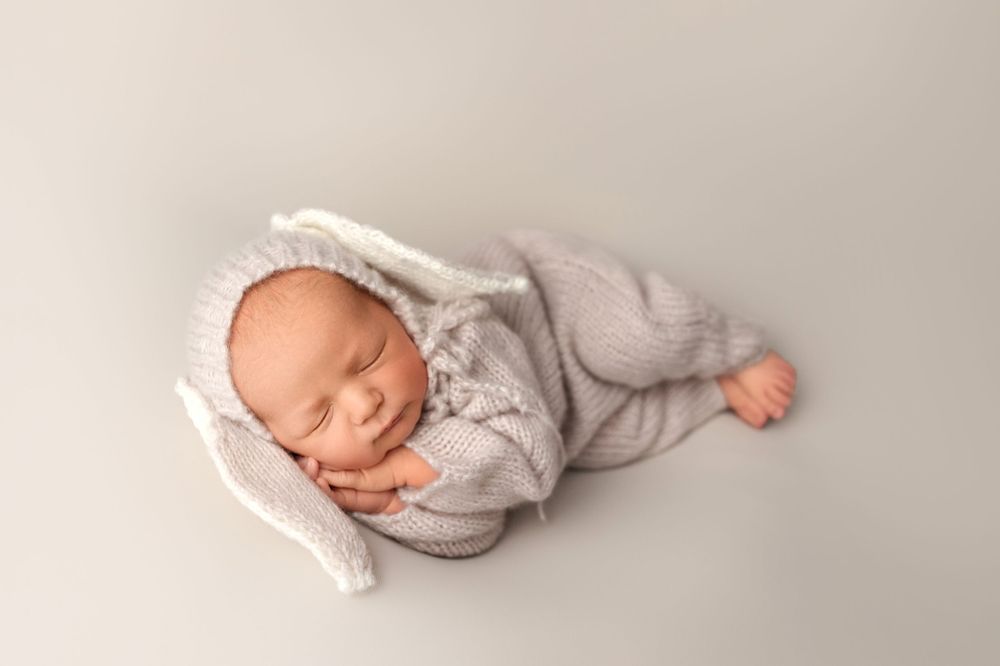 Sleeping baby in grey outfit and white bonnet on neutral background.