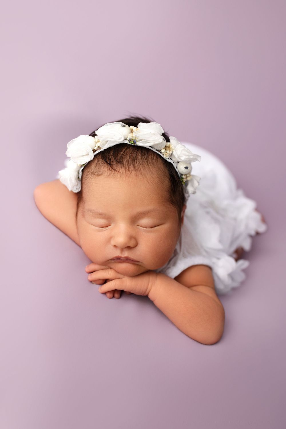 Newborn in white outfit and floral crown sleeping on purple background.