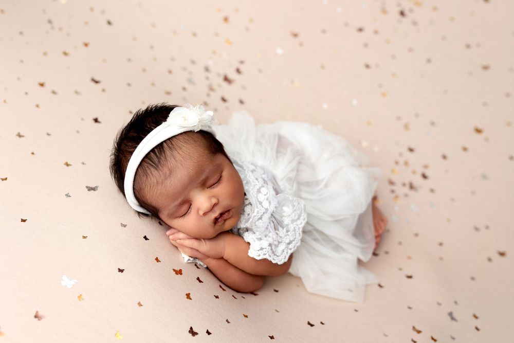 Newborn baby sleeping peacefully in white lace dress and headband on beige backdrop with gold confetti.