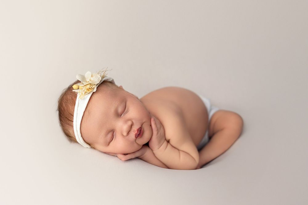 Peaceful sleeping newborn baby wearing delicate floral headband curled up on soft beige backdrop.