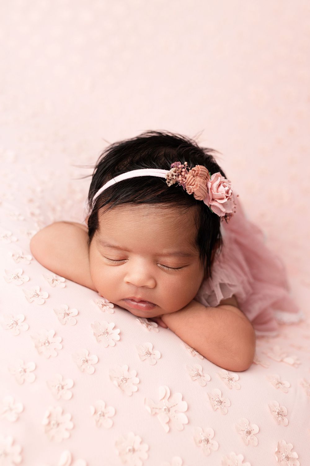 Sleeping newborn with floral headband rests on pink textured fabric.