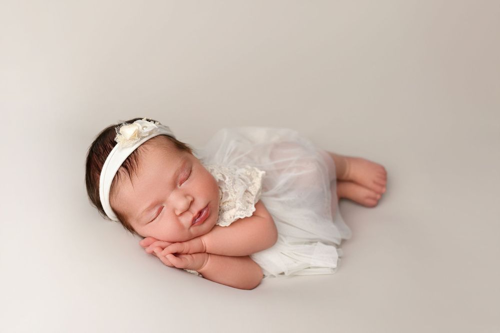 Sleeping newborn in white lace dress and headband lying on soft white backdrop during professional photo shoot.