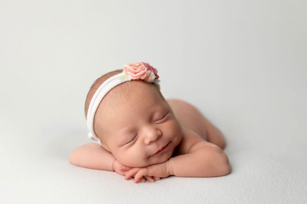 Smiling newborn baby girl wearing a pink floral headband, peacefully sleeping on a white blanket.
