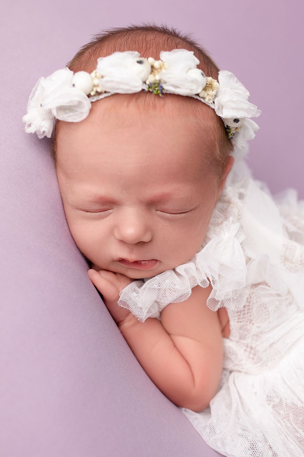 Sleeping newborn wearing white lace dress and floral crown against purple background.